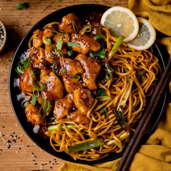 A plate of Lemon Pepper Chicken with sesame seeds and spring onions, served with stir-fried noodles, lemon slices, and chopsticks.