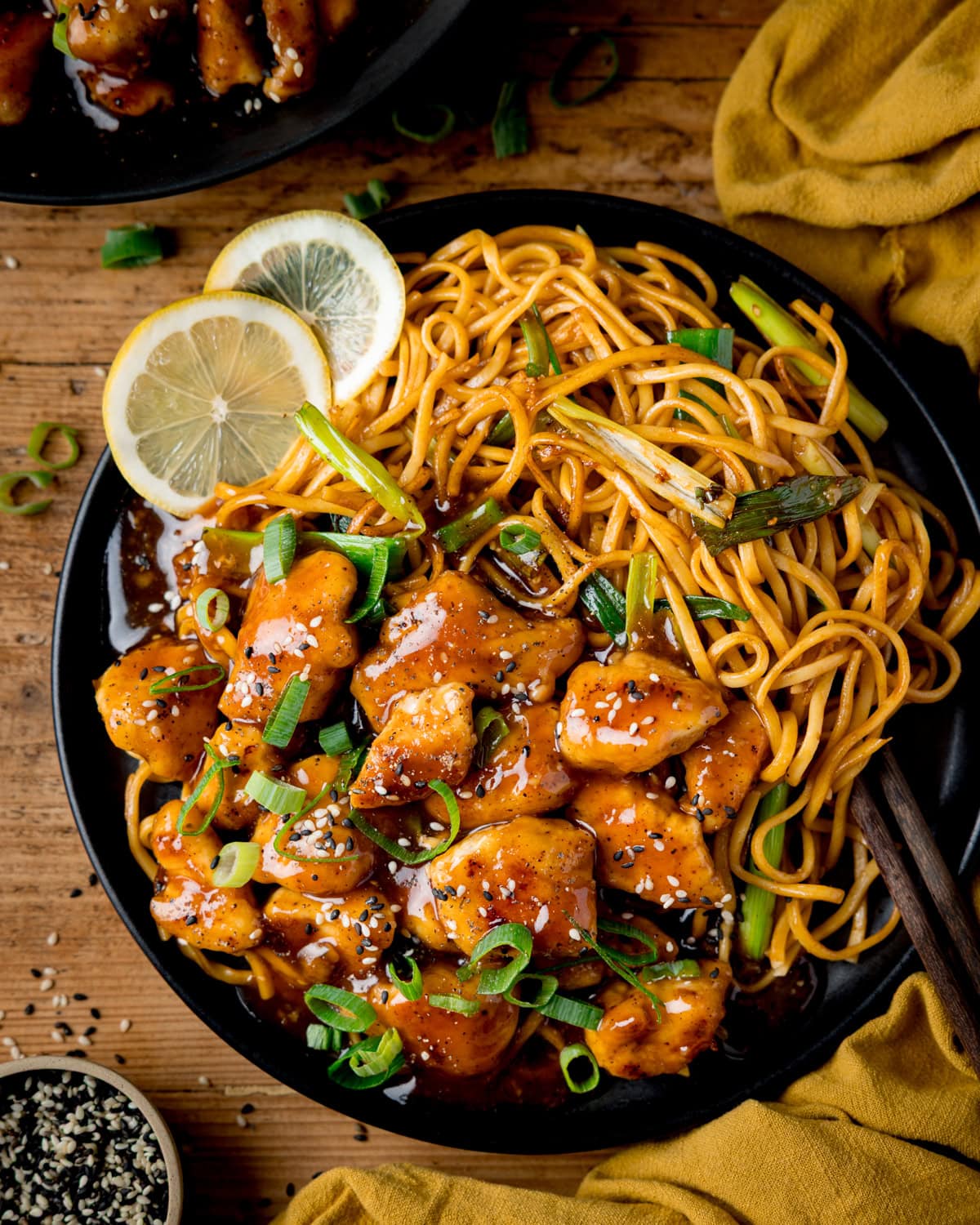 A plate of saucy Lemon Pepper Chicken pieces with sliced spring onions and sesame seeds, served with stir-fried noodles, lemon slices, and chopsticks on a wooden table.