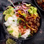 A minced beef fajita bowl with white rice, grated cheese, seasoned minced beef with peppers, sliced red onions, avocado, lime wedges, soured cream, salsa, and coriander.