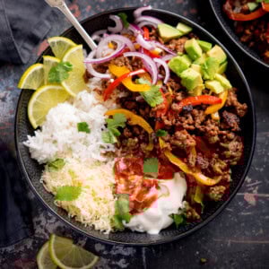 A minced beef fajita bowl with white rice, grated cheese, seasoned minced beef with peppers, sliced red onions, avocado, lime wedges, soured cream, salsa, and coriander.