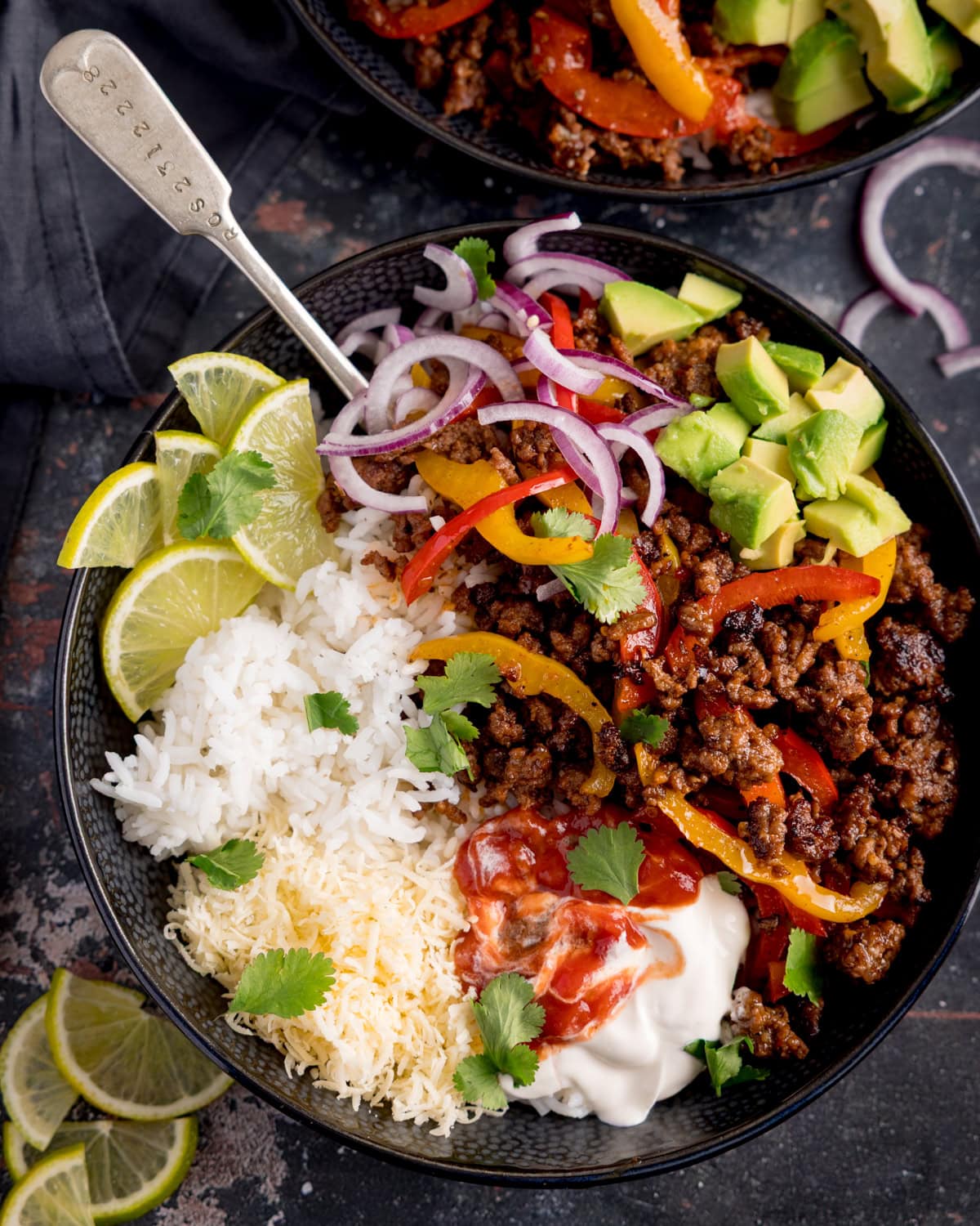 A minced beef fajita bowl with white rice, grated cheese, sliced red onion, peppers, avocado, lime wedges, soured cream, salsa and coriander, served with a spoon.