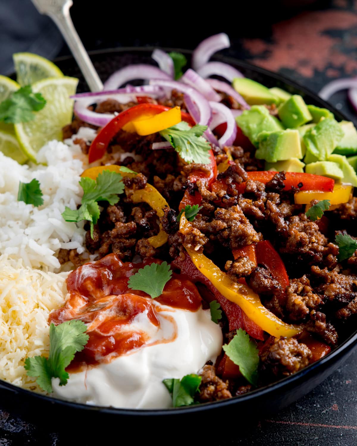A close up of mince beef fajita bowl filled with seasoned minced beef, sliced peppers, white rice, avocado, grated cheese, soured cream, salsa, red onion, lime wedges, and fresh coriander.