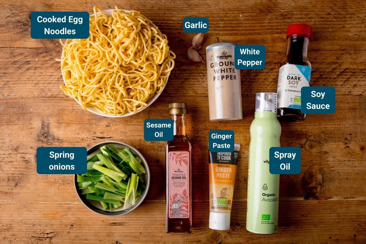 Ingredients for a fried noodle side dish on a wooden surface: cooked egg noodles, spring onions, garlic, white pepper, soy sauce, sesame oil, ginger paste, and spray oil.