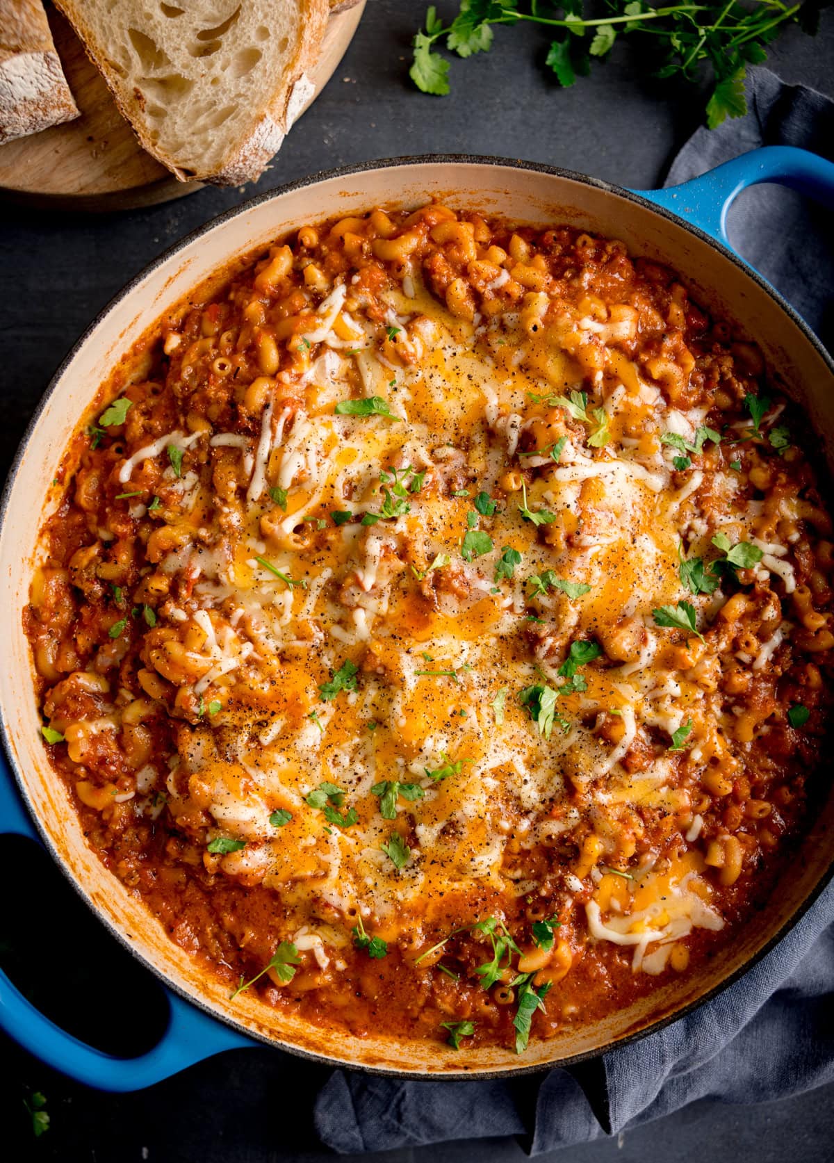 A casserole pan filled with minced beef mac n cheese, topped with melted cheese and chopped herbs, with slices of bread and parsley nearby.