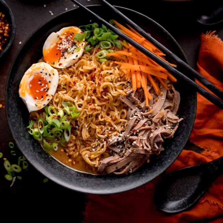 A black bowl of Peanut Butter Ramen with sliced beef, soft-boiled eggs, spring onions, shredded carrots, and sesame seeds, with chopsticks resting on the rim.