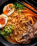 A close up overhead image of a bowl of peanut butter ramen topped with sliced beef meat, chopped spring onions (scallions) julienned carrots, a halved soft-boiled egg, sesame seeds, and chopsticks resting on the edge.