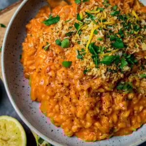A plate of creamy tomato risotto topped with chopped parsley, lemon zest, crispy breadcrumbs, and black pepper, served with a fresh lemon wedge on the side.