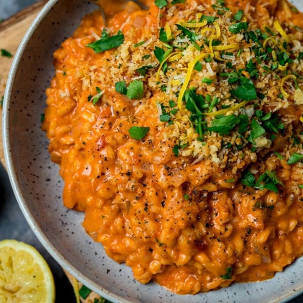 A plate of creamy tomato risotto topped with chopped parsley, lemon zest, crispy breadcrumbs, and black pepper, served with a fresh lemon wedge on the side.