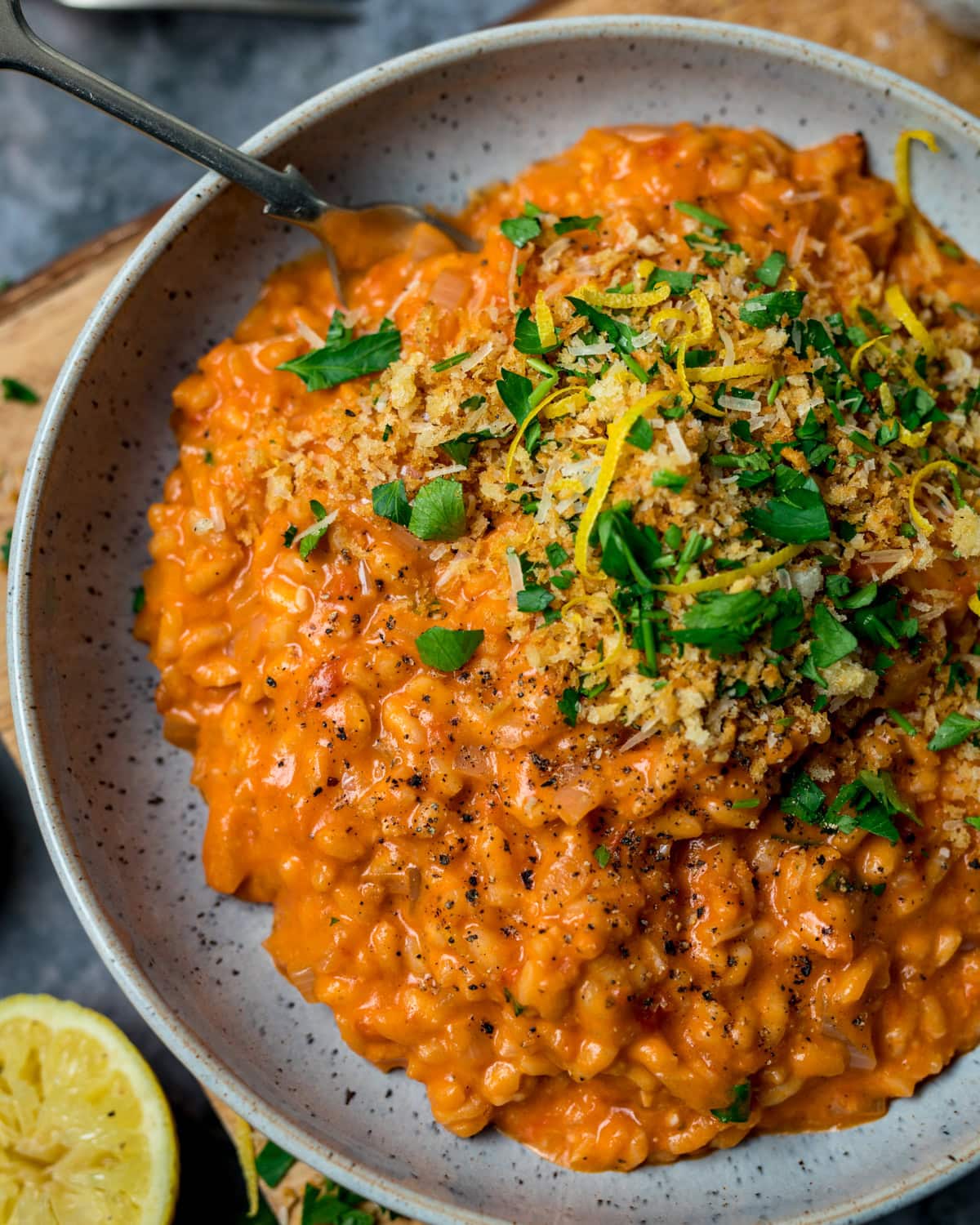 A bowl of creamy tomato risotto topped with crispy breadcrumbs, chopped parsley, lemon zest, and cracked black pepper, with a spoon and a sliced lemon nearby.