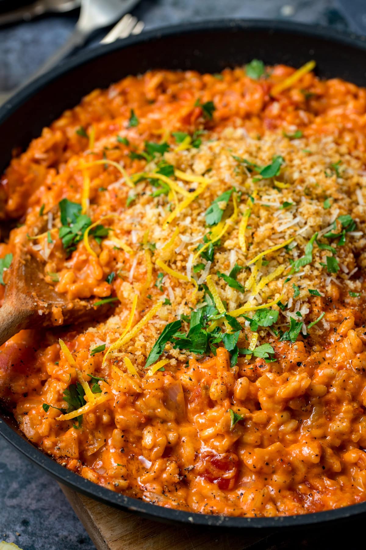 A frying pan filled with creamy tomato risotto, topped with breadcrumbs, lemon zest and parsley, with a wooden spoon on the side.
