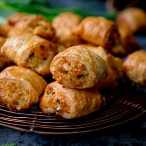 A close-up of golden-brown vegetarian sausage rolls stacked on a wire cooling rack, with a blurred background of more rolls and green herbs.