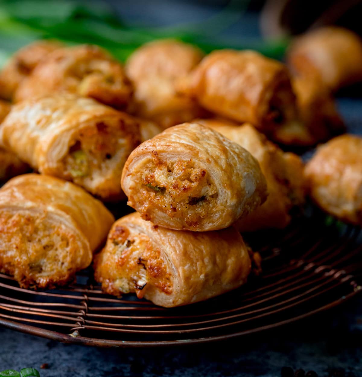 A close-up of several golden-brown meat free sausage rolls stacked on a wire cooling tray, with a blurred background.