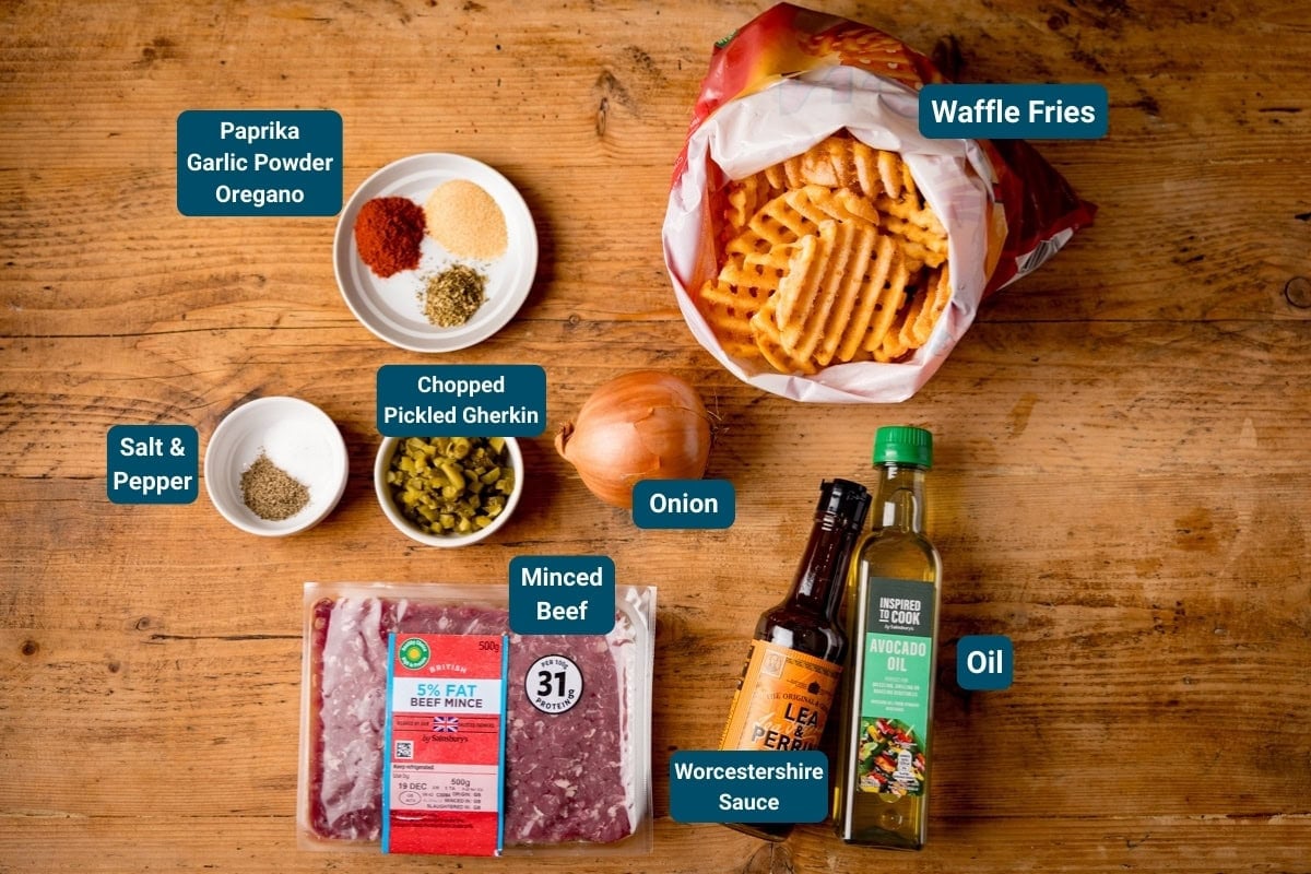 A wooden table with ingredients labeled: waffle fries, onion, oil, Worcestershire sauce, minced beef, chopped pickled gherkin, salt and pepper, paprika, garlic powder, and oregano.