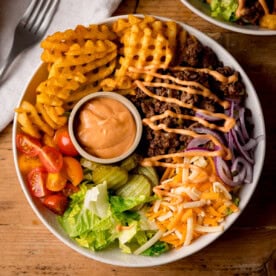 A burger bowl with waffle chips, minced beef, grated cheese, sliced red onions, lettuce, gherkins, cherry tomatoes, and a small pot of sauce, drizzled with dressing.