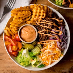 A burger bowl with waffle chips, minced beef, grated cheese, sliced red onions, lettuce, gherkins, cherry tomatoes, and a small pot of sauce, drizzled with dressing.