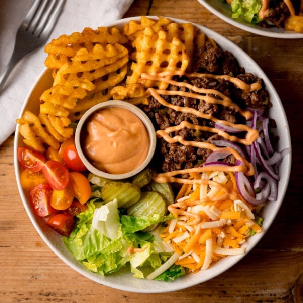 A burger bowl with waffle chips, minced beef, grated cheese, sliced red onions, lettuce, gherkins, cherry tomatoes, and a small pot of sauce, drizzled with dressing.