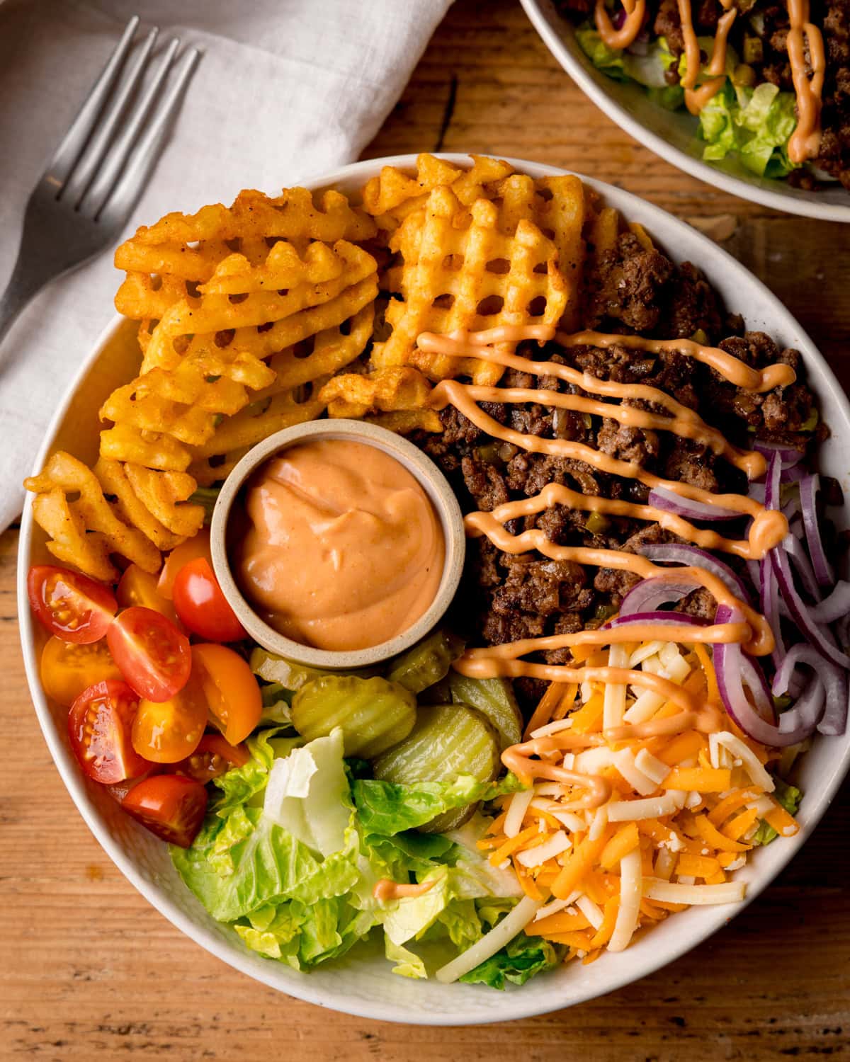 A burger bowl with waffle chips, seasoned ground beef drizzled with burger sauce, grated cheddar, lettuce, cherry tomatoes, gherkins, red onions, and a small cup of burger sauce on a wooden table.