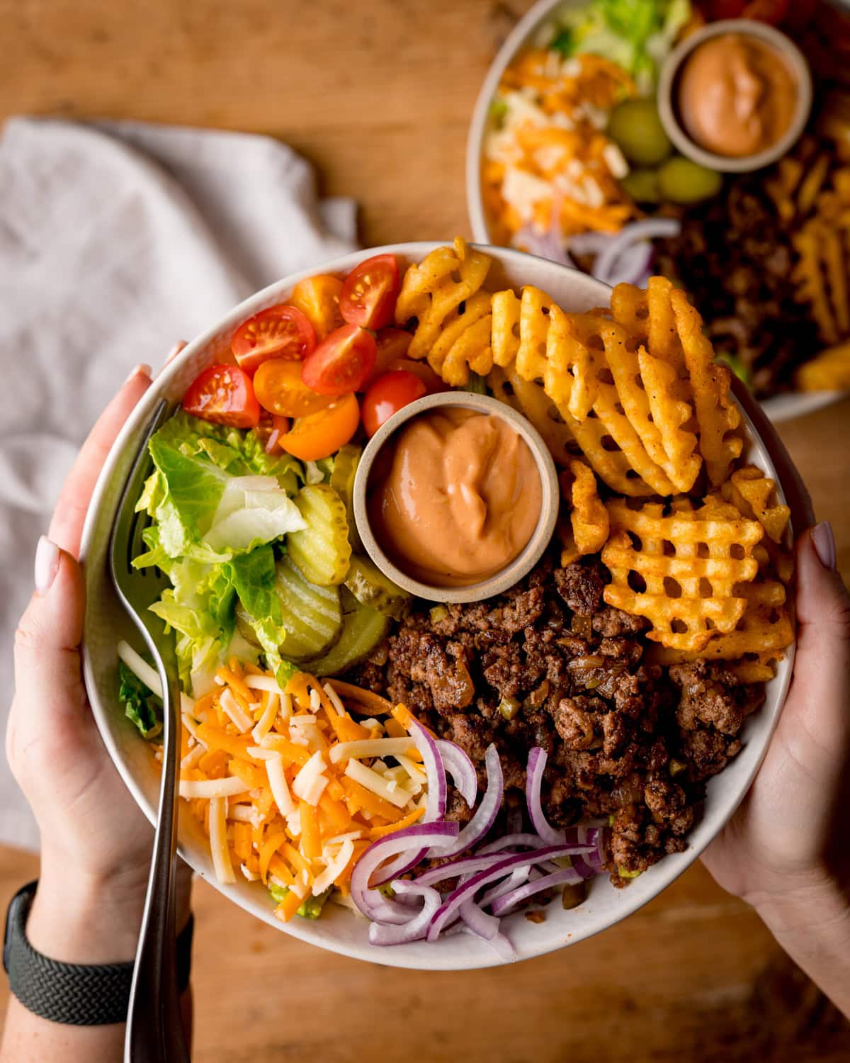 A burger bowl with waffle chips, minced beef, grated cheese, lettuce, cherry tomatoes, gherkins, red onions, and a pot of burger sauce held in two hands.