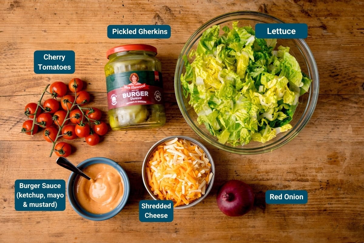 A wooden table with labelled ingredients: cherry tomatoes, pickled gherkins, lettuce, burger sauce, grated cheese, and a red onion.