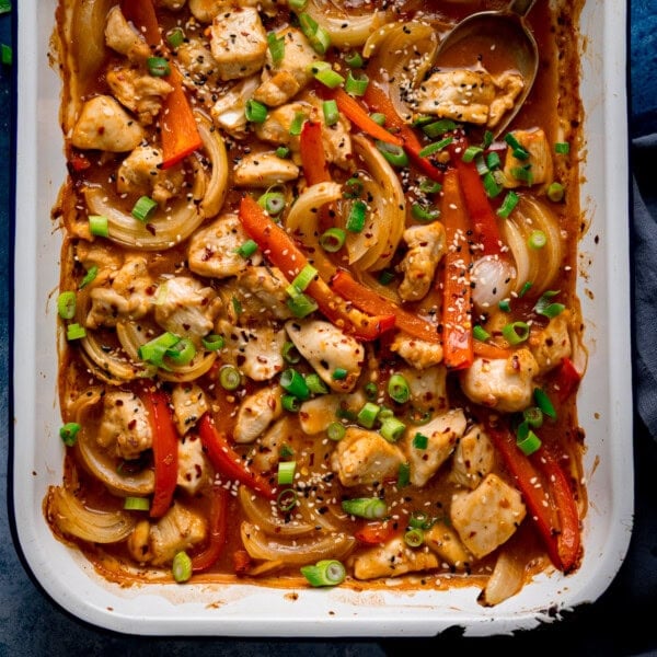 Overhead of a chicken 'stir fry' traybake with diced chicken, sliced red peppers, onions, spring onions, sesame seeds and sauce in a white dish.