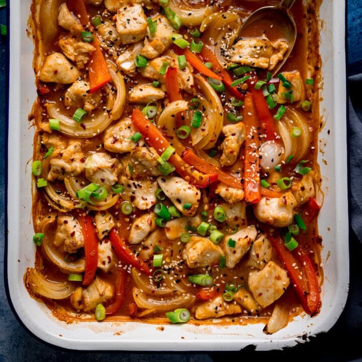 Overhead of a chicken 'stir fry' traybake with diced chicken, sliced red peppers, onions, spring onions, sesame seeds and sauce in a white dish.