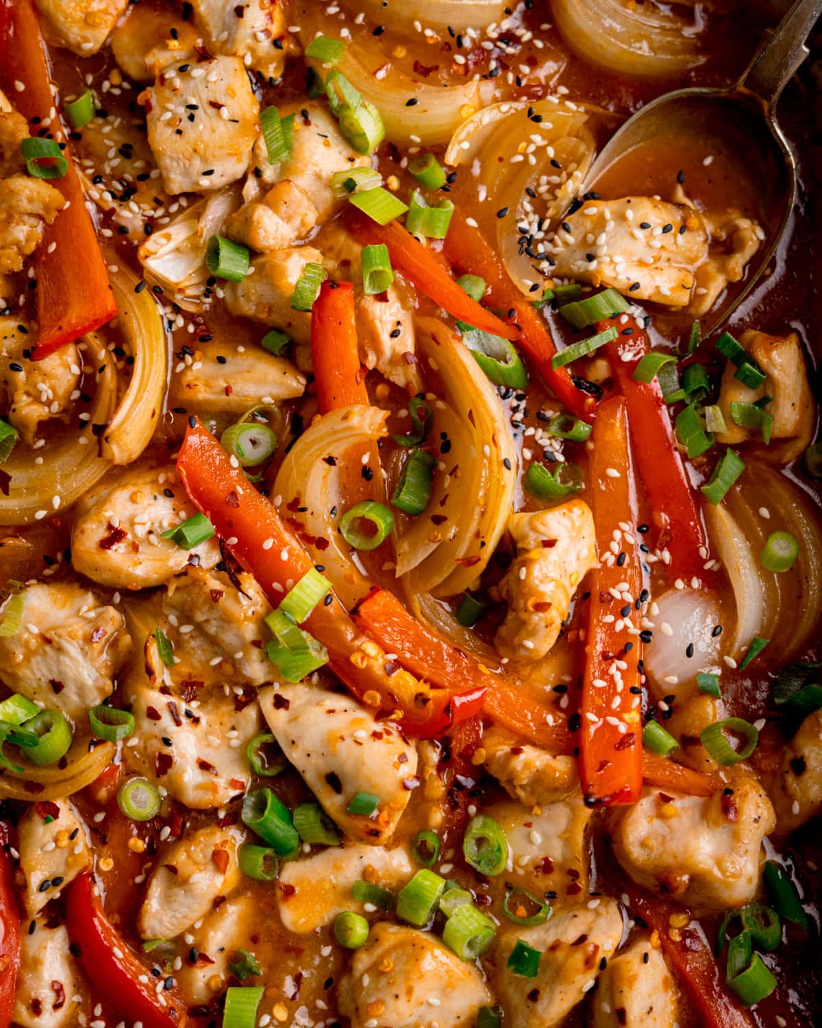 Close up overhead shot of cooked chicken pieces with red pepper, onion slices, and spring onions in a stir-fry sauce, topped with sesame seeds.