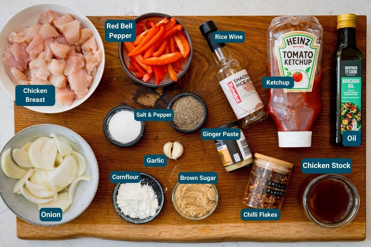 A wooden board displays labelled ingredients for a stir fry chicken tray bake: chicken breast, onion, red pepper, seasonings, sauces, cornflour, brown sugar, and oil. Ingredients are labelled.