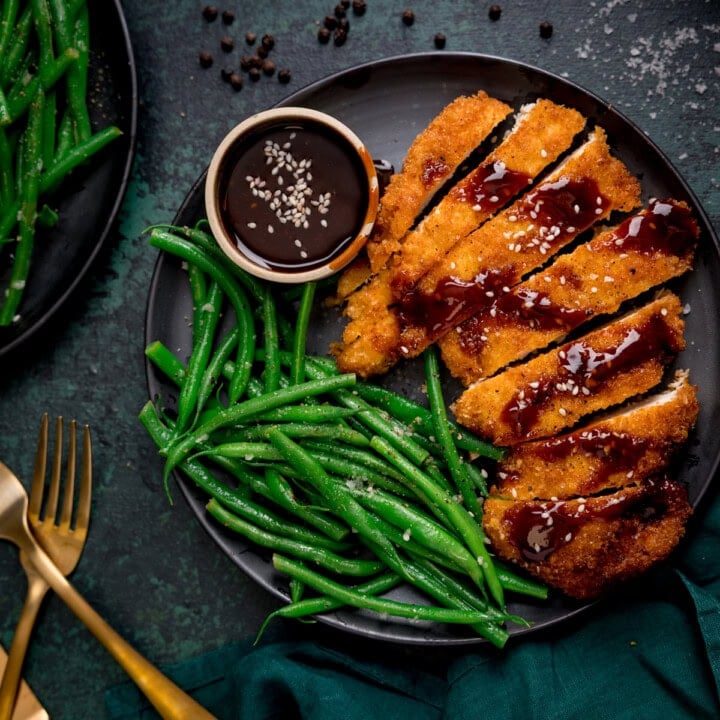 A plate of breaded chicken cutlets drizzled with a gochujang sauce, served with green beans and a small bowl of extra sauce, with a gold fork and knife on the side.