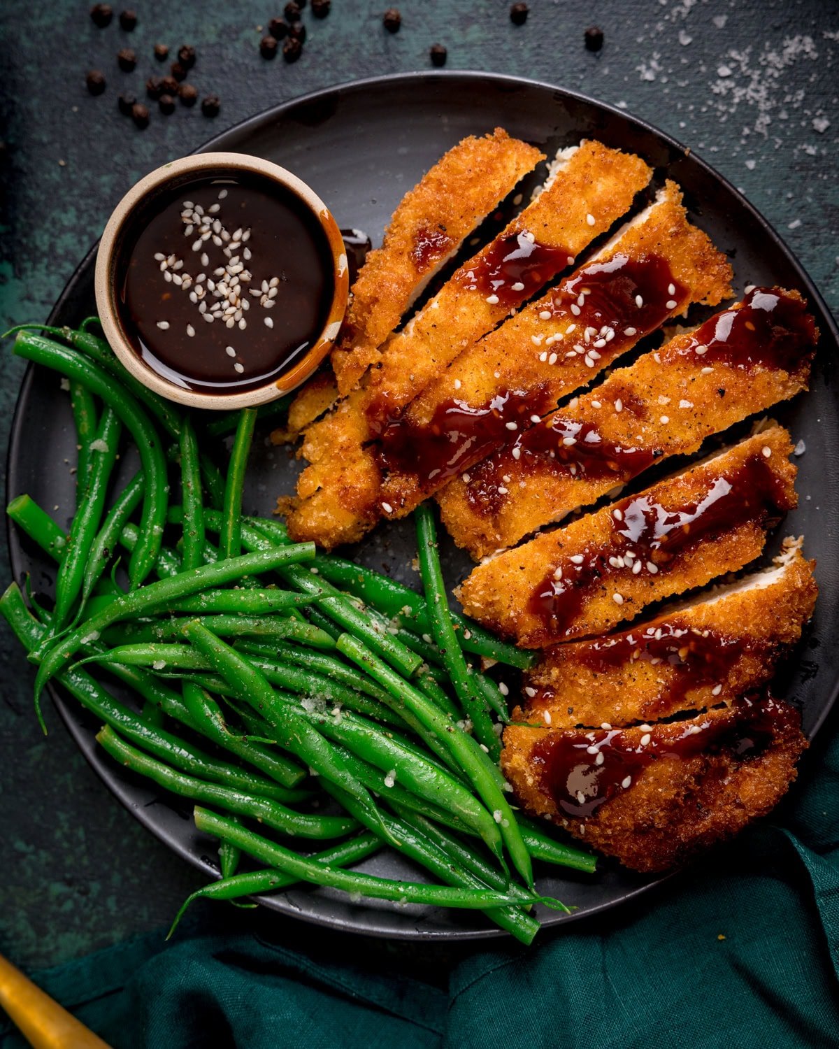 A plate with sliced crispy chicken cutlets topped with a Korean inspired sauce and sesame seeds, served with green beans and a small bowl of dark dipping sauce.