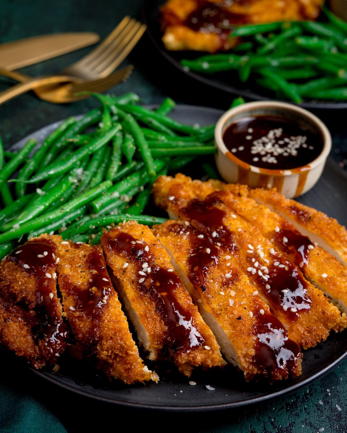 A plate of breaded and sliced chicken cutlets topped with a sweet sticky Korean sauce, served with green beans and a small pot of dipping sauce, garnished with sesame seeds.