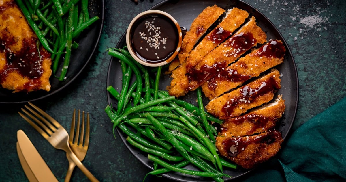 A plate with sliced breaded chicken cutlets topped with gochujang sauce, steamed green beans, a small bowl of dipping sauce, and gold cutlery on the side.
