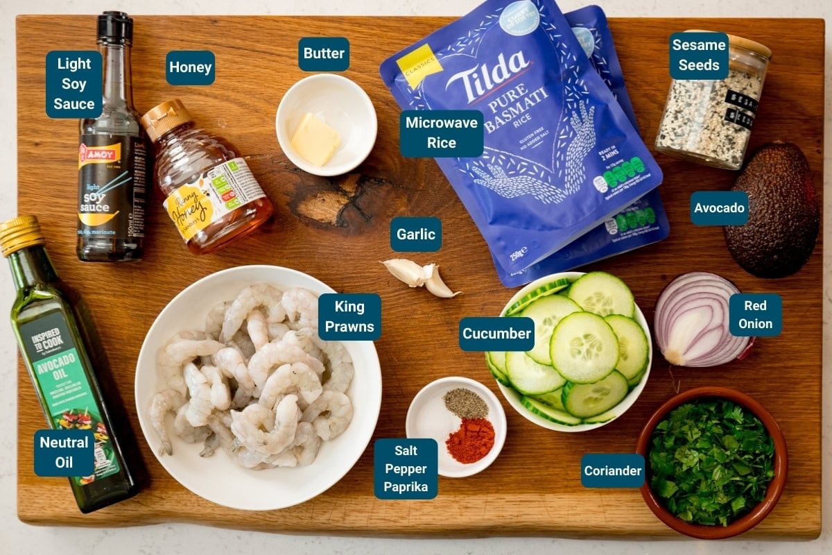 A wooden board topped with labelled ingredients for honey garlic prawn rice bowls: king prawns, rice, butter, oil, soy sauce, honey, garlic, cucumber, paprika, salt, pepper, coriander, onion, avocado and sesame seeds.