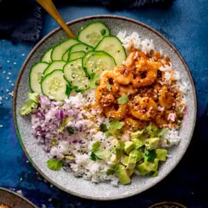 A bowl of white rice topped with cooked honey garlic prawns, sliced cucumber, chopped red onion, diced avocado, fresh coriander, and sesame seeds.