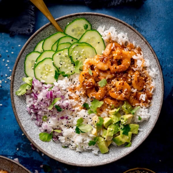 A bowl of white rice topped with cooked honey garlic prawns, sliced cucumber, chopped red onion, diced avocado, fresh coriander, and sesame seeds.