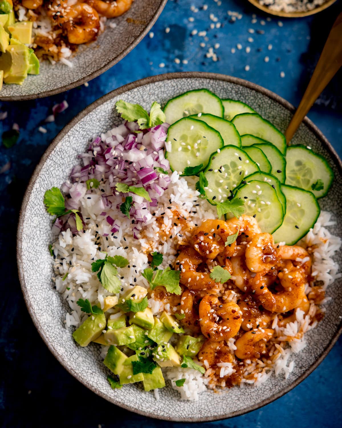 A bowl with white rice, cooked honey garlic prawns in sauce, sliced cucumber, diced red onion, avocado chunks, coriander, and sesame seeds on a blue background.