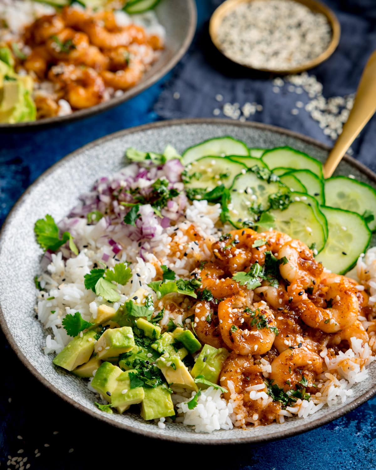 A bowl with white rice, cooked prawns in honey garlic sauce, diced avocado, chopped red onion, cucumber slices, coriander, and sesame seeds on top.