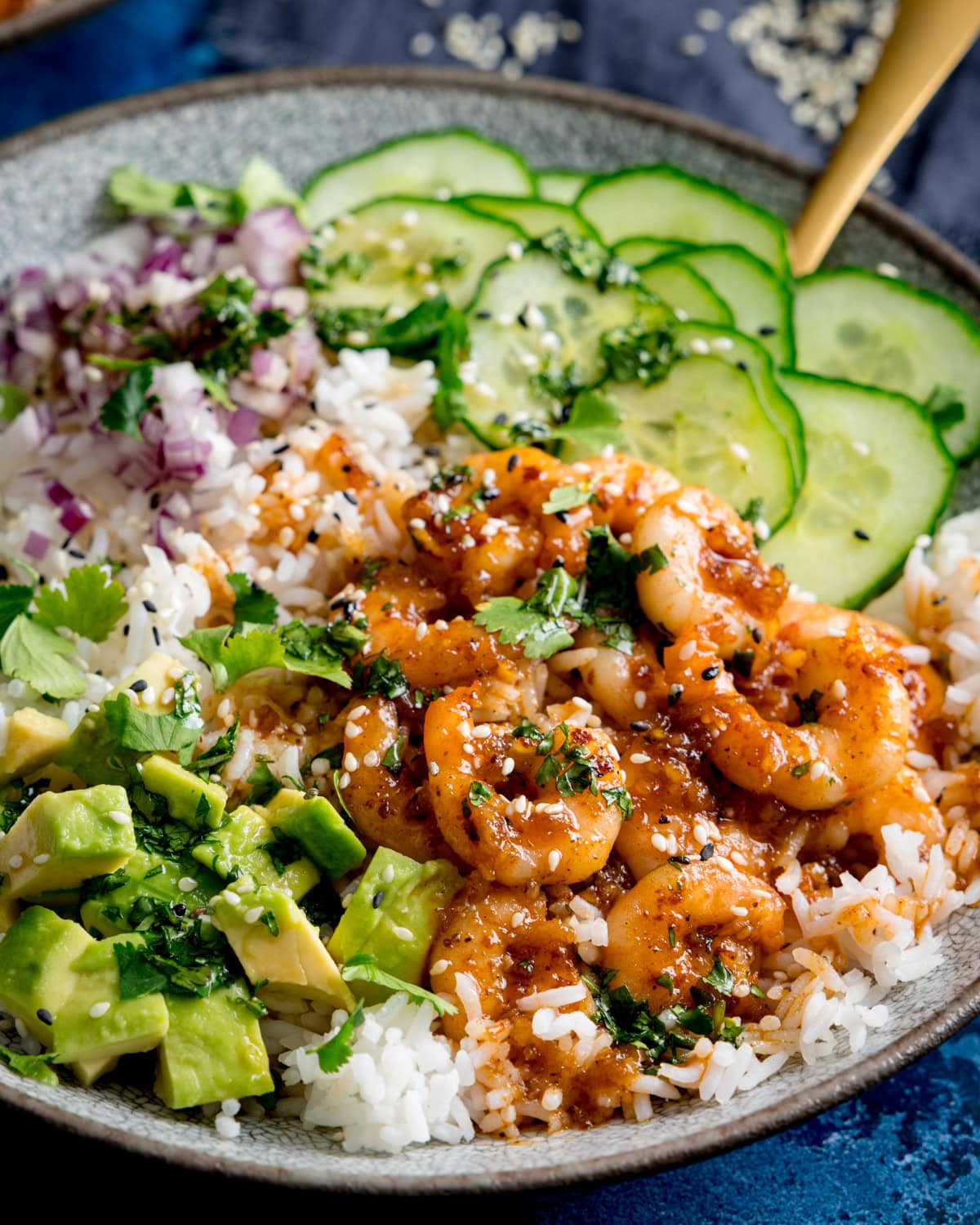 A close up of a honey garlic prawn rice bowl with; white rice topped with honey glazed prawns, sliced cucumber, diced avocado, chopped red onion, coriander, and sesame seeds.