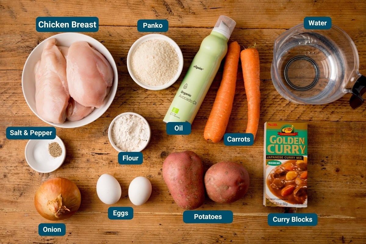 Top-down view of chicken katsu curry ingredients: chicken breasts, panko, oil, carrots, potatoes, curry blocks, onion, eggs, flour, salt, pepper and water on a wooden table. Each item is labelled.