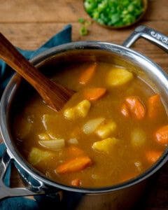 A pan of Japanese curry sauce with potatoes, carrots, and onions being stirred with a wooden spoon. Chopped spring onions are in a small dish behind the pan.