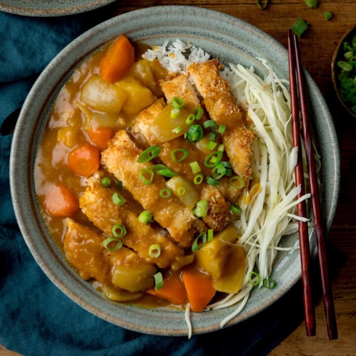A bowl of Chicken Katsu Curry with rice, breaded chicken fillets, sliced carrots and potatoes, shredded cabbage, and chopped spring onions, served with chopsticks.