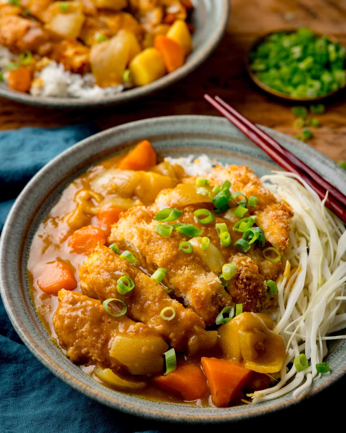 A bowl of Japanese Katsu Curry rice topped with a crispy chicken cutlet, sliced spring onions and shredded cabbage, served with chopsticks on the side. A further bowl of curry is in the background, next to a small dish of spring onions.