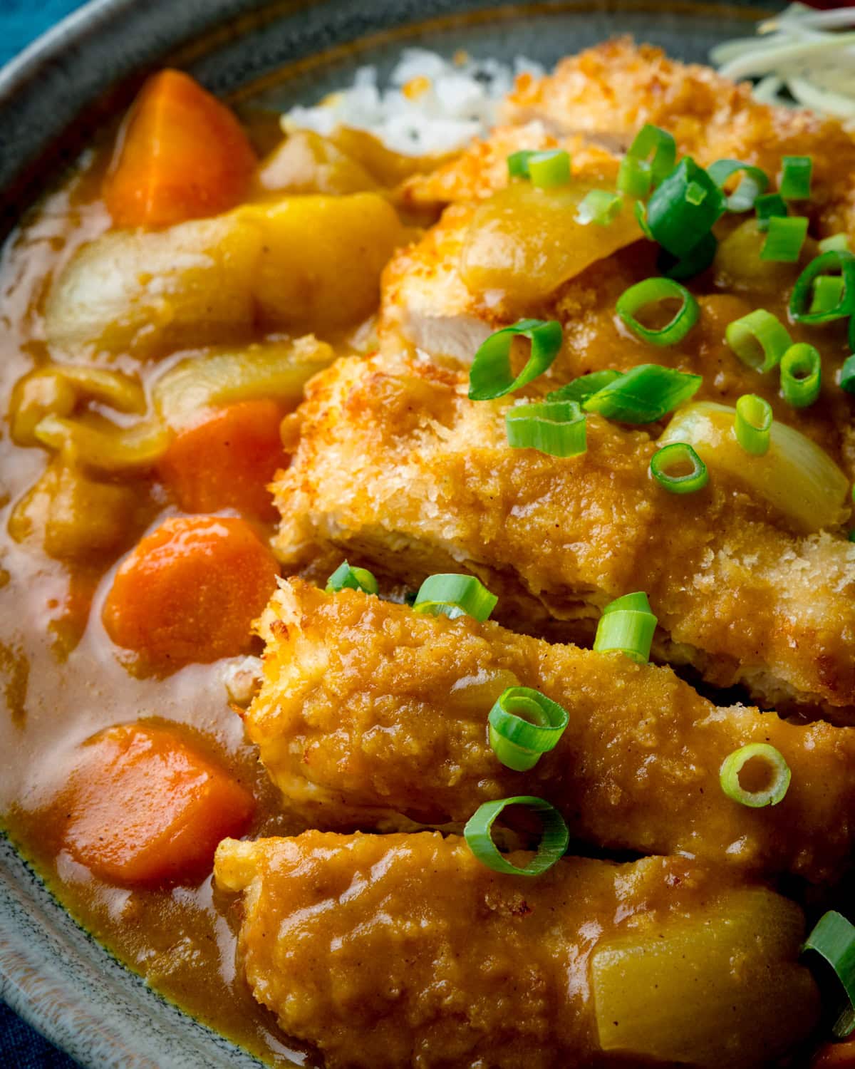 A close up image of a bowl of Chicken Katsu Curry with rice, chopped spring onions, carrots and potatoes.