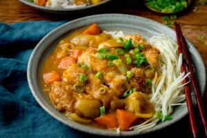 Wide image of Chicken Katsu Curry in a green bowl with rice and shredded cabbage. The chicken is topped with spring onions. A set of chopsticks rests on the bowl. The bowl is on a teal napkin.