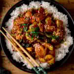 A dark bowl of white rice topped with saucy mongolian chicken, spring onions, and sesame seeds, with a pair of wooden chopsticks on the side.