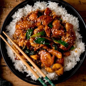 A dark bowl of white rice topped with saucy mongolian chicken, spring onions, and sesame seeds, with a pair of wooden chopsticks on the side.
