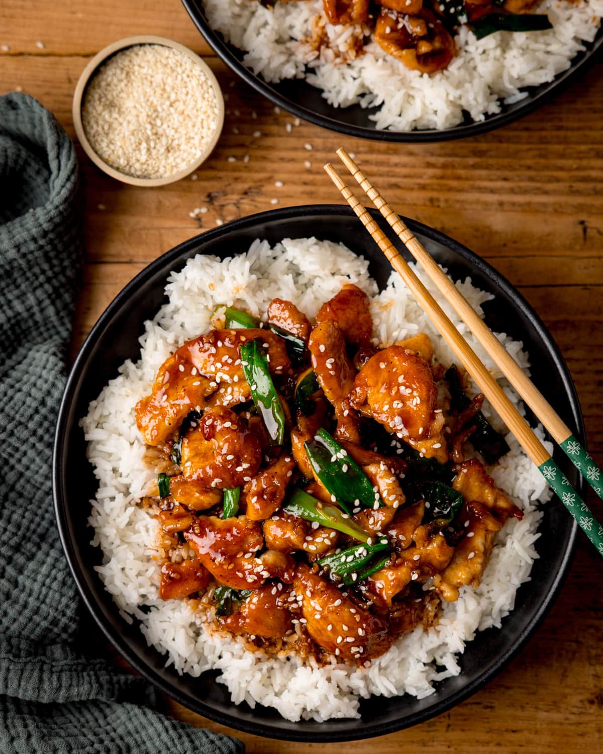 A bowl of white rice topped with Mongolian chicken, spring onions, and sesame seeds, with chopsticks on the side and a small bowl of sesame seeds nearby.