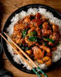 A black bowl filled with white rice topped with sticky takeout style mongolian chicken, spring onions, sesame seeds, and a pair of chopsticks resting on the side.