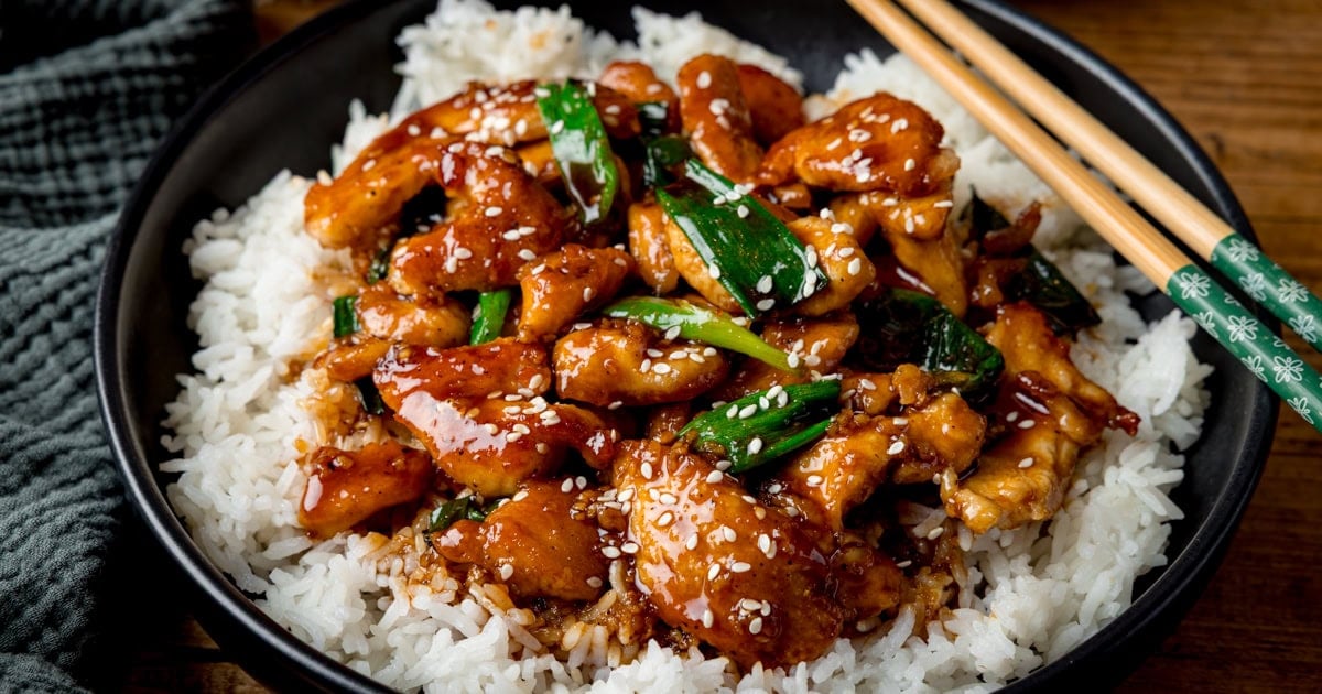 A bowl of white rice topped with sticky mongolian chicken, spring onions, sesame seeds, and sauce, with chopsticks resting on the bowl&rsquo;s edge.