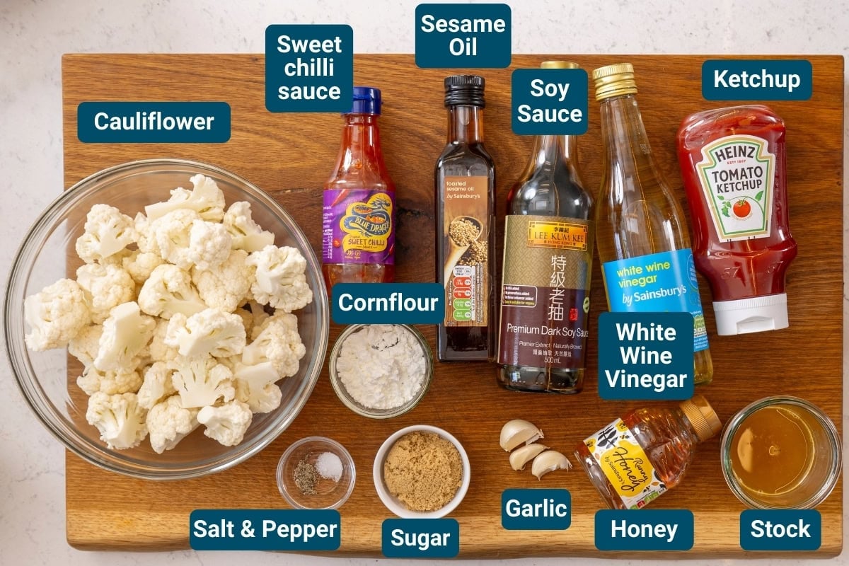 A wooden board displays cauliflower florets in a bowl, surrounded by labelled bottles and bowls of sauces, seasonings, and condiments, including soy sauce, tomato sauce, and vinegar.