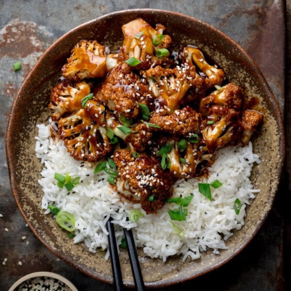 A bowl of white rice topped with saucy sesame cauliflower, garnished with chopped spring onions, with black chopsticks on the side.